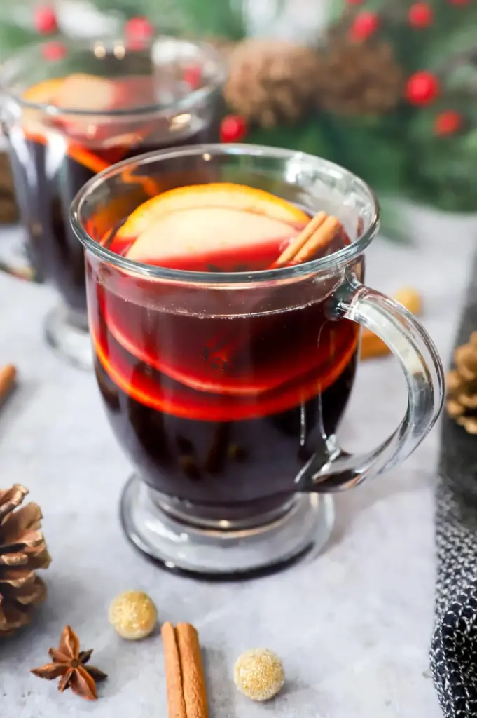 Mulled wine in a clear glass on a white table garnished with apples and cinnamon sticks